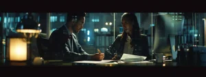 two agents discussing paperwork at a desk with a computer displaying a government website.